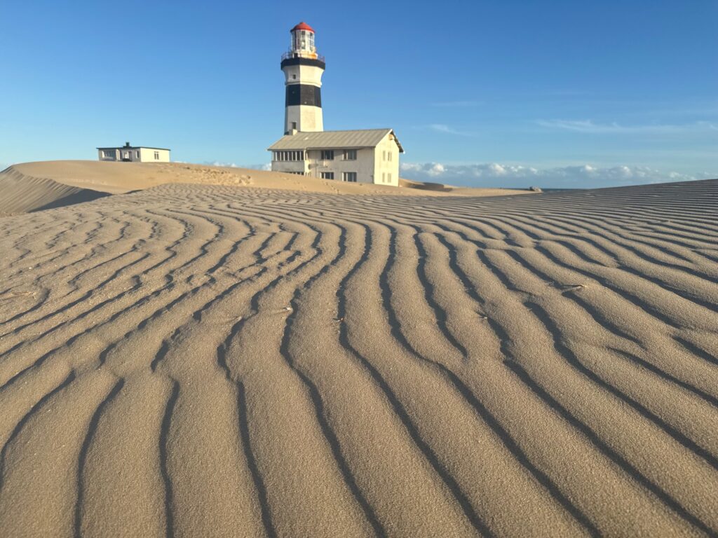 Sanddüne mit wellenförmigen Mustern im Vordergrund und Leuchtturm mit angrenzendem Gebäude im Hintergrund unter blauem Himmel