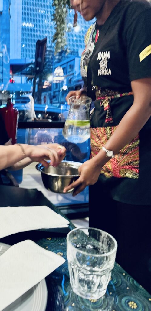 Person mit schwarzem T-Shirt und gemusterter Schürze hält eine Karaffe mit Wasser und eine Metallschüssel, während eine andere Person darin die Hände spült