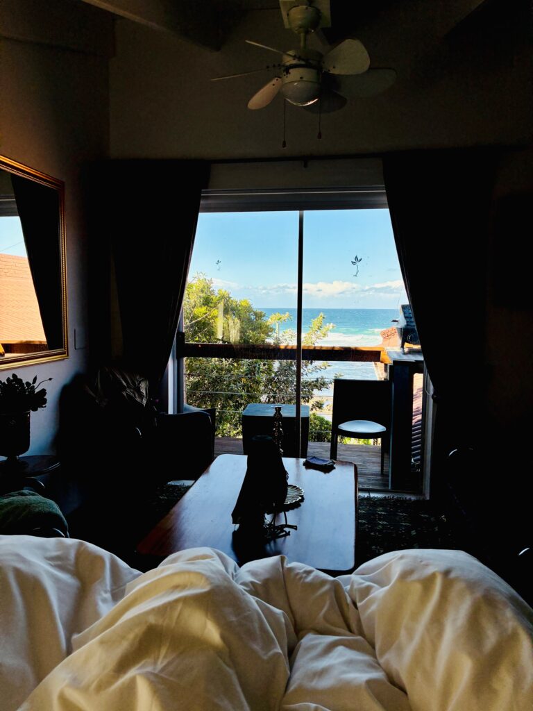 Blick aus einem Zimmer mit Bett auf eine Terrasse und das Meer im Hintergrund bei Tageslicht