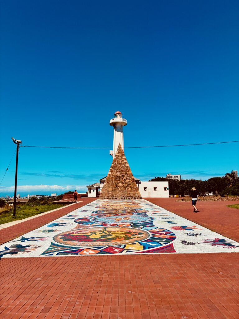 Leuchtturm mit pyramidenförmigem Steinsockel und farbiger Mosaikfläche davor auf rotem Pflaster unter blauem Himmel