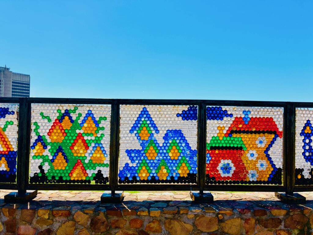 Bunte Mosaikbilder aus Plastikflaschendeckeln, die an einem Geländer befestigt sind, mit Mustern aus Dreiecken und einem Haus