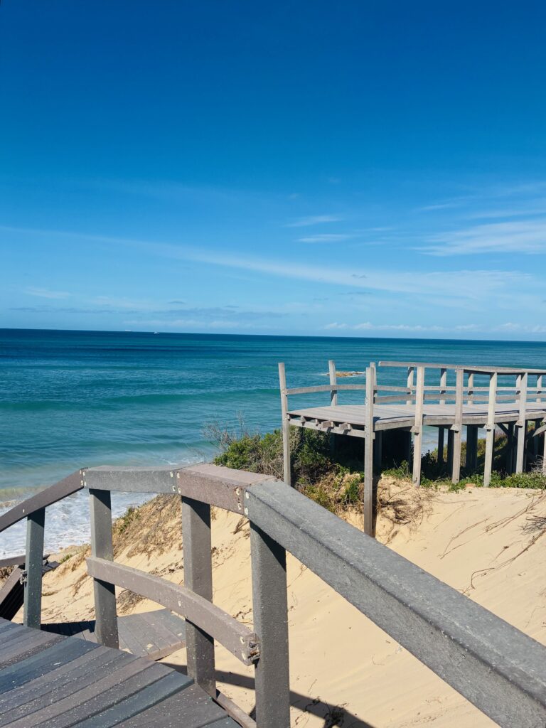 Abrupt in der Luft endender Holzsteg mit Geländer über Sanddünen am Meer unter blauem Himmel