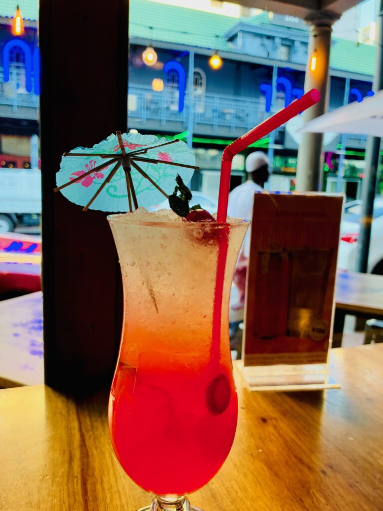 Cocktailglas mit rotem Getränk, rotem Trinkhalm und kleinem Papierschirm auf Holztisch vor Fenster mit unscharfem Hintergrund, in dem ein Schwarzer Mann mit weißer Kopfbedeckung vorbeigeht