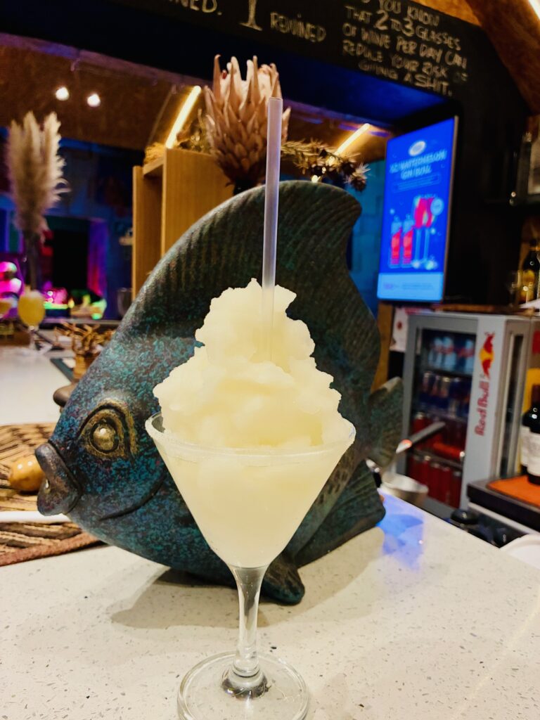 Cocktailglas mit einem gefrorenen, schneeweißen Getränk und einem Strohhalm vor einer großen, fischförmigen Dekoration auf einer Bar.