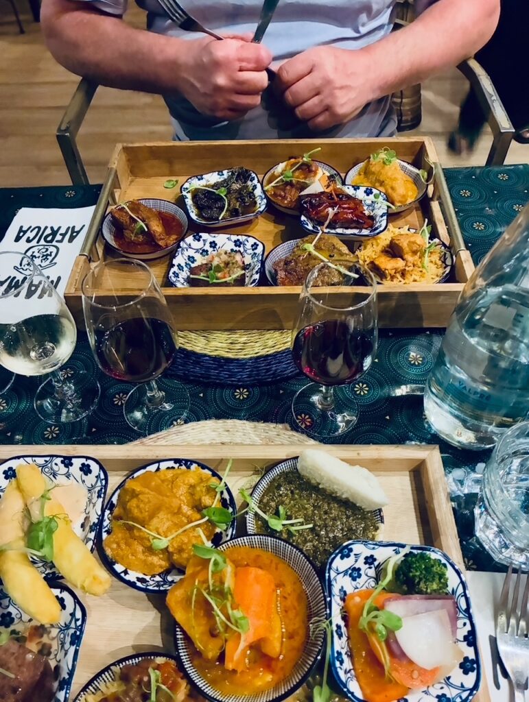 Zwei Tabletts mit verschiedenen kleinen Gerichten und Gläsern mit Rot- und Weißwein auf einem Tisch, im Hintergrund eine Person mit Besteck in den Händen