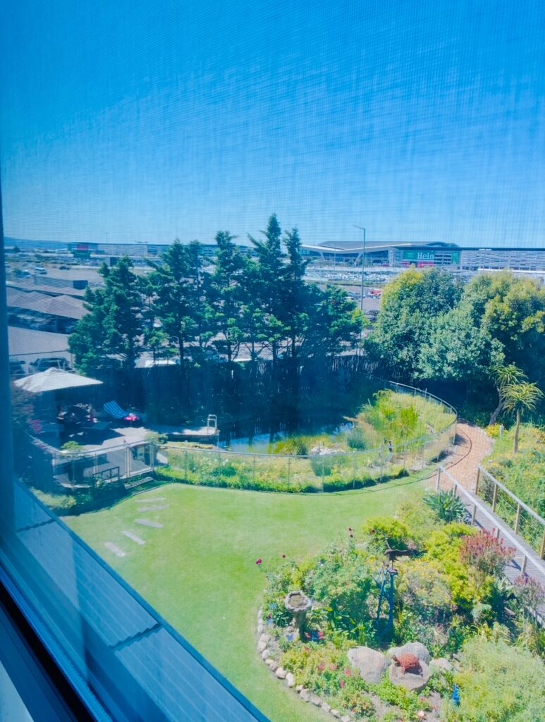 Blick aus einem Fenster auf einen Garten mit Rasen, Blumenbeeten, Büschen und einem kleinen Teich, im Hintergrund Bäume und das Terminal des Flughafens Kapstadt