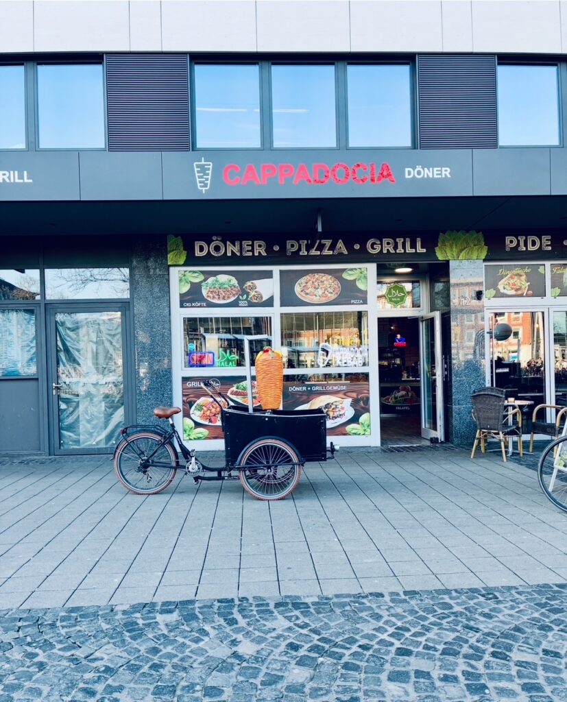 Lastenfahrrad mit großem Döner-Spieß aus Plastik vor dem Imbiss Cappadocia Döner in Münster mit Schaufensteranzeigen für Döner, Pizza und Grillgerichte