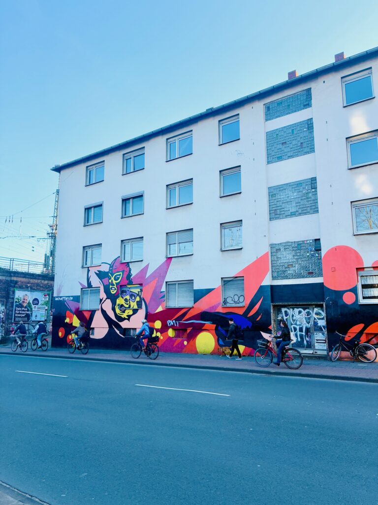 Mehrstöckiges Wohnhaus in Münster mit bunter Graffiti-Wand und mehreren Radfahrern auf der Straße davor unter blauem Himmel