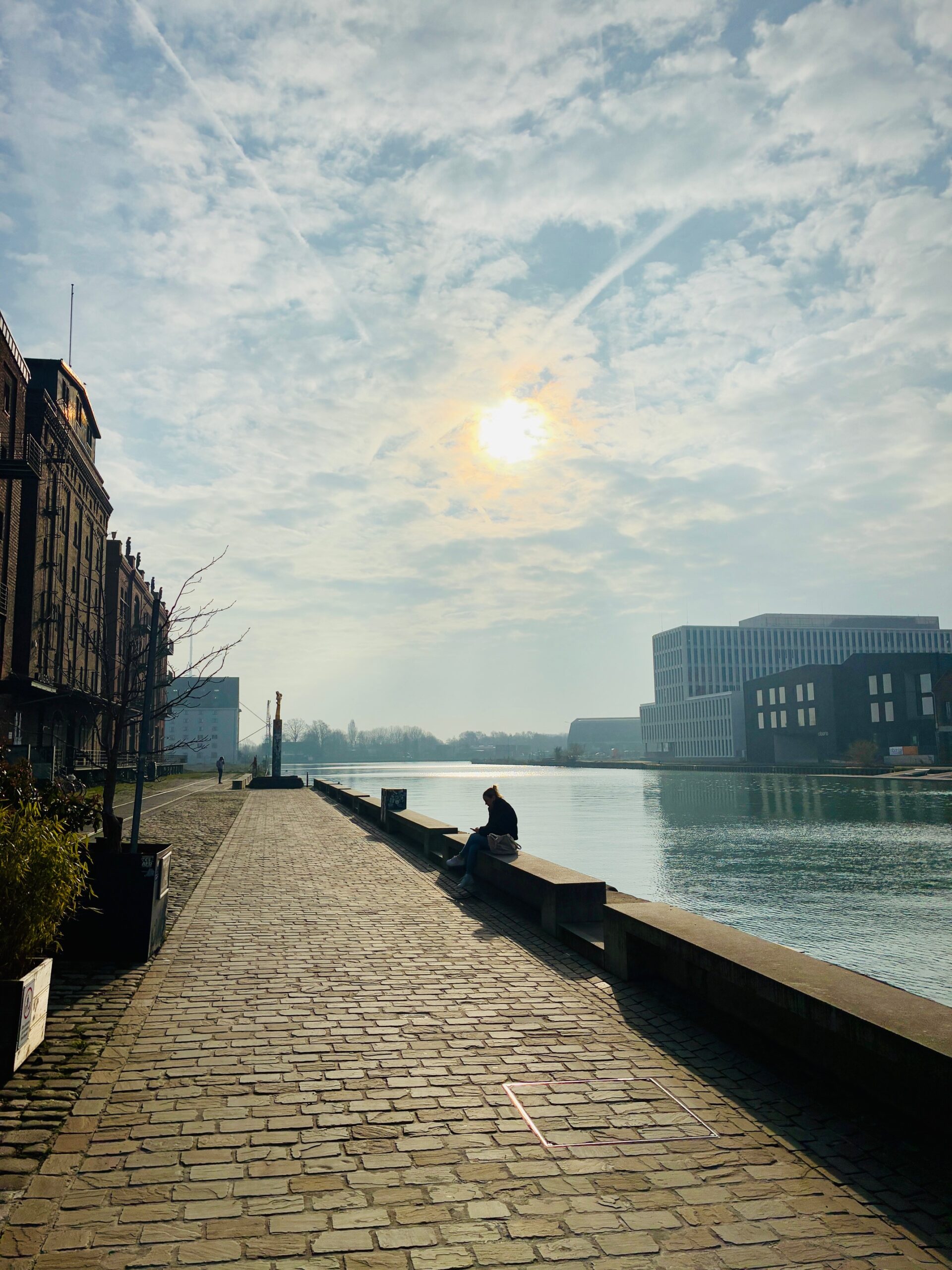 Kopfsteinpflasterweg am Hafen in Münster mit einer Person im Gegenlicht, die auf einer niedrigen Mauer am Wasser sitzt, im Hintergrund moderne Gebäude und Himmel mit Sonne und freundlichen Wolken