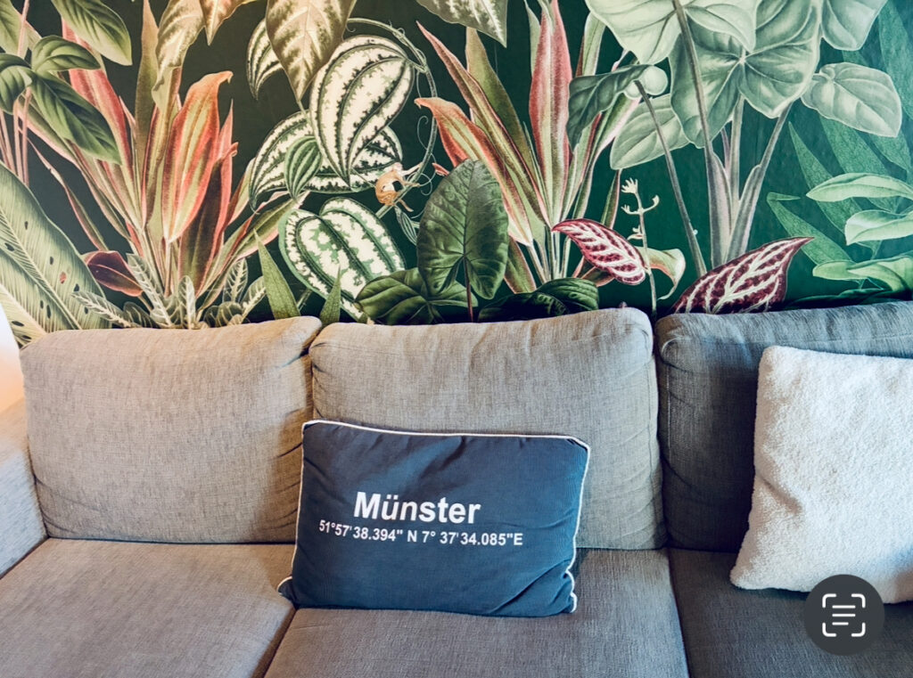Hellgraues Sofa mit einem dunkelblauen Kissen mit der Aufschrift 'Münster' und den Koordinaten der Stadt vor einer Wand mit großem, farbigem Pflanzenmuster