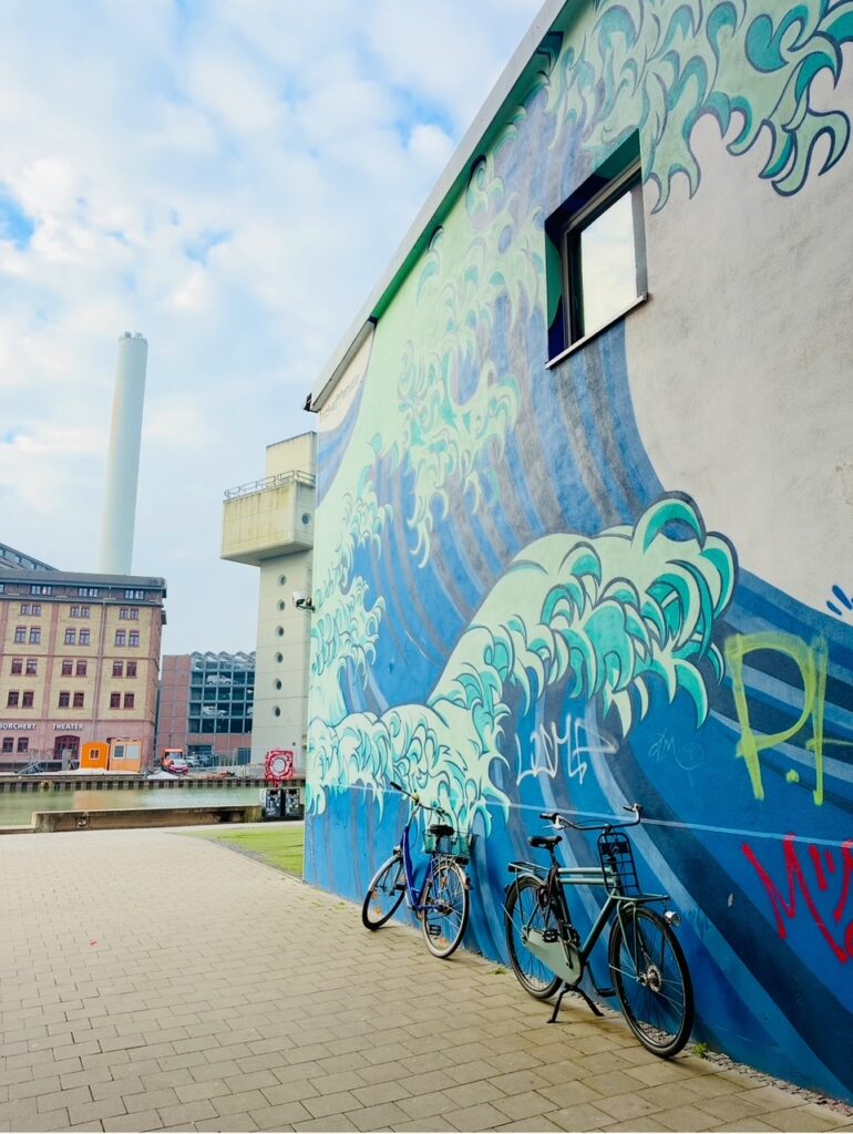 Fahrräder lehnen an einer Hauswand mit großflächigem Wellen-Graffiti am Hafen in Münster, im Hintergrund Industriegebäude und ein hoher Schornstein