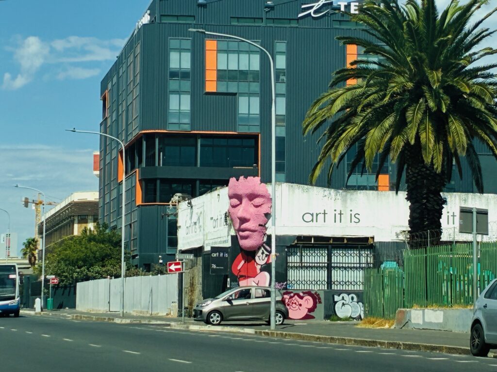 Straßenszene mit moderner Gebäudefassade, Palmen, Graffiti und einem silbernen Auto am Straßenrand und Eckgebäude mit einem großen, rosa Reliefgesicht sowie dem Schriftzug 'art it is'