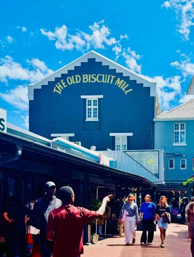 Menschen gehen und stehen vor einem Gebäude mit der Aufschrift 'THE OLD BISCUIT MILL' unter einem blauen Himmel mit freundlichen Wolken