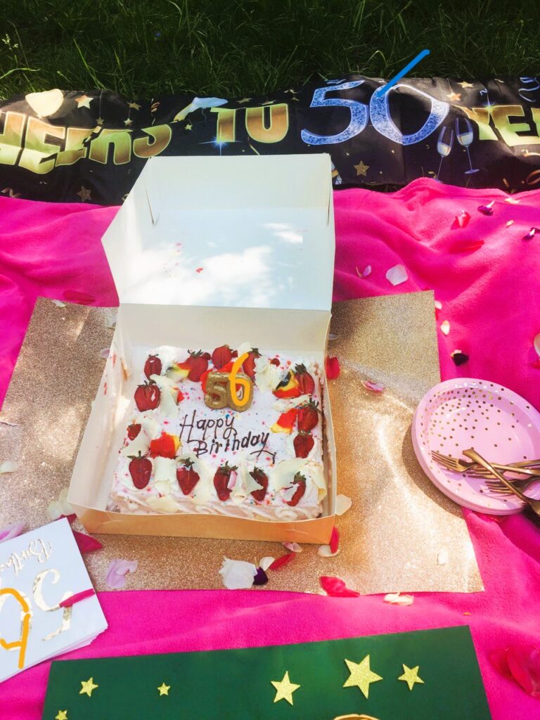 Geburtstagstorte mit Erdbeeren und einer gelben Kerze in Form der Zahl 50, die von Hand in 56 geändert wurde, auf einer pinken Picknickdecke mit goldfarbenen Tellern und Besteck
