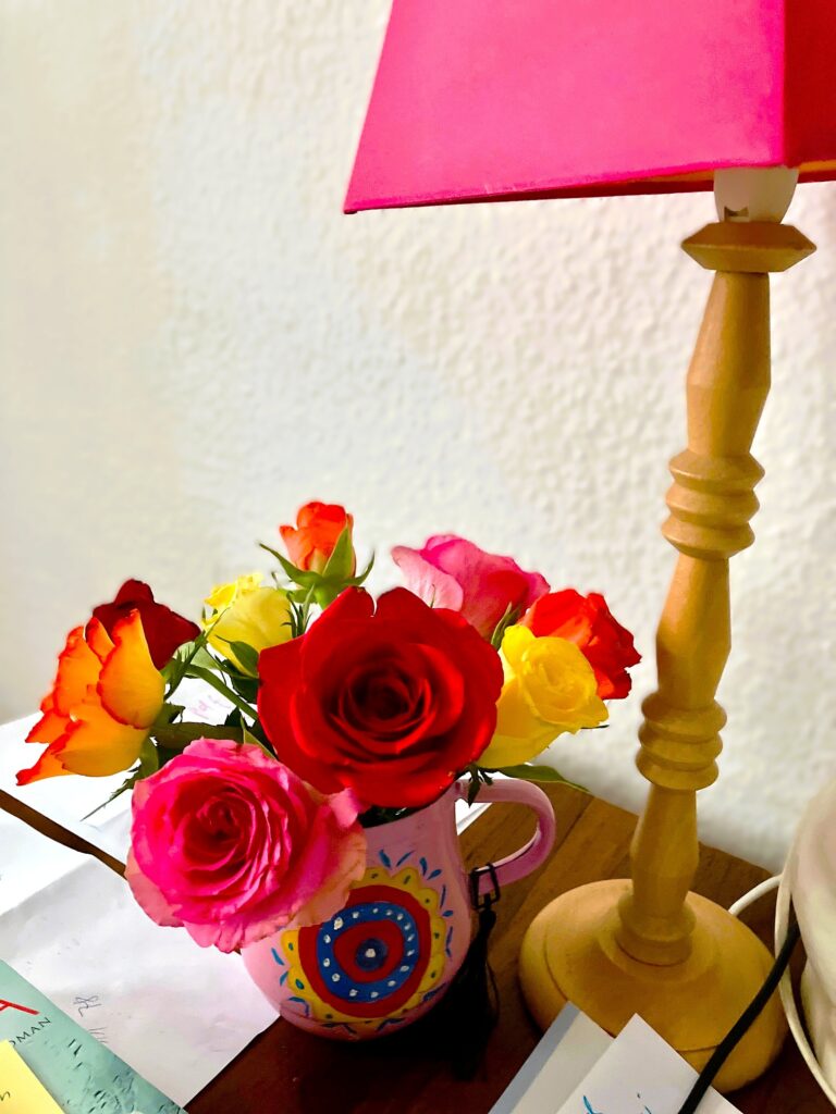 Bunte Rosen in einer bemalten Tasse neben einem Holzlampenfuß mit rotem Lampenschirm auf einem Tisch