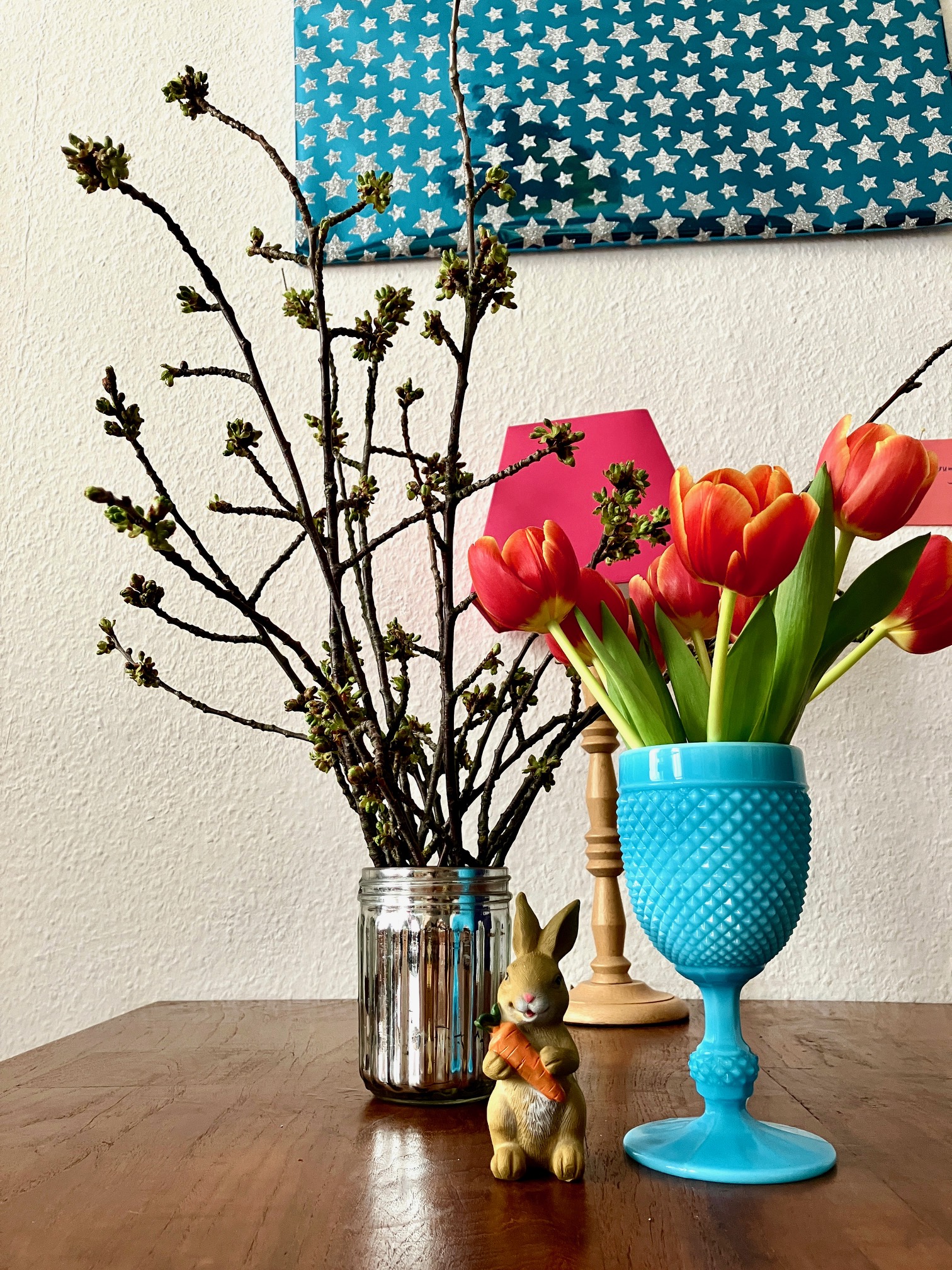 Zweig mit Knospen in Glasvase, daneben orange Tulpen in blauer Vase und kleine Hasenfigur auf Holztisch