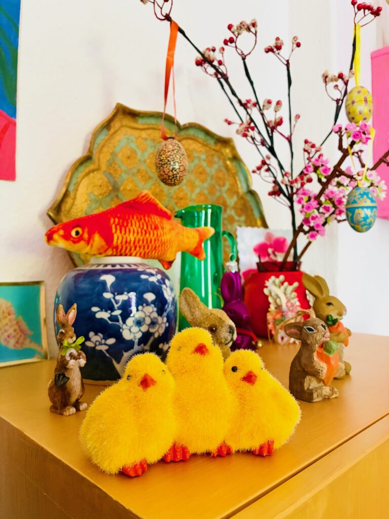 Farbenfrohe Osterdekoration auf einem Sideboard mit drei gelben Osterküken aus Plüsch vor Osterhasenfiguren, einer blauen Vase mit Blumenmustern, Zweigen mit hängenden bemalten Eiern und weiteren kleinen Hasenfiguren sowie einer japanischen Vase mit einem bunten Fisch