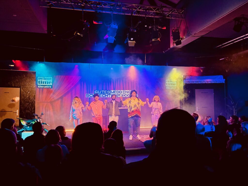 Fünf Schauspieler auf einer Bühne mit buntem Bühnenlicht vor Publikum in einem Theater