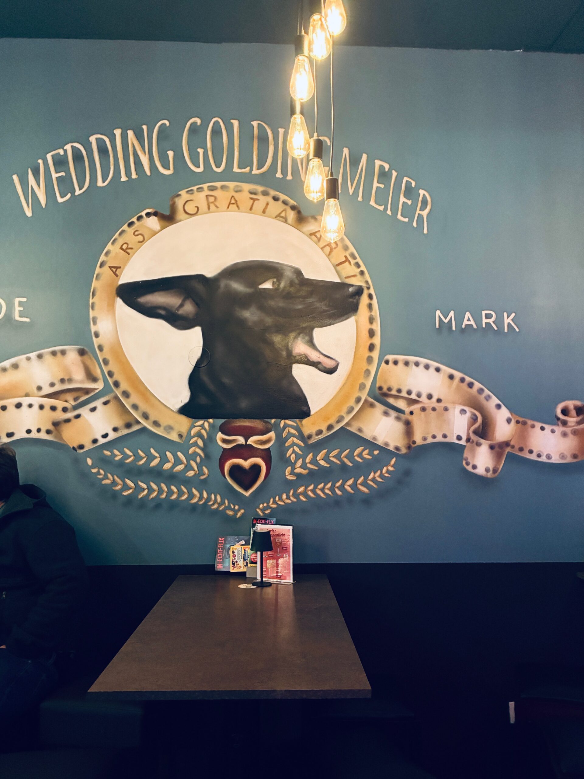 Wandgemälde mit einem schwarzen Hundekopf in einem goldenen Kreis, umgeben von Schriftzügen und einem stilisierten Band, davor ein Tisch mit Menükarten. Das Gemälde ist dem Logo der Filmfirma Metro Goldwyn Mayer nachempfunden