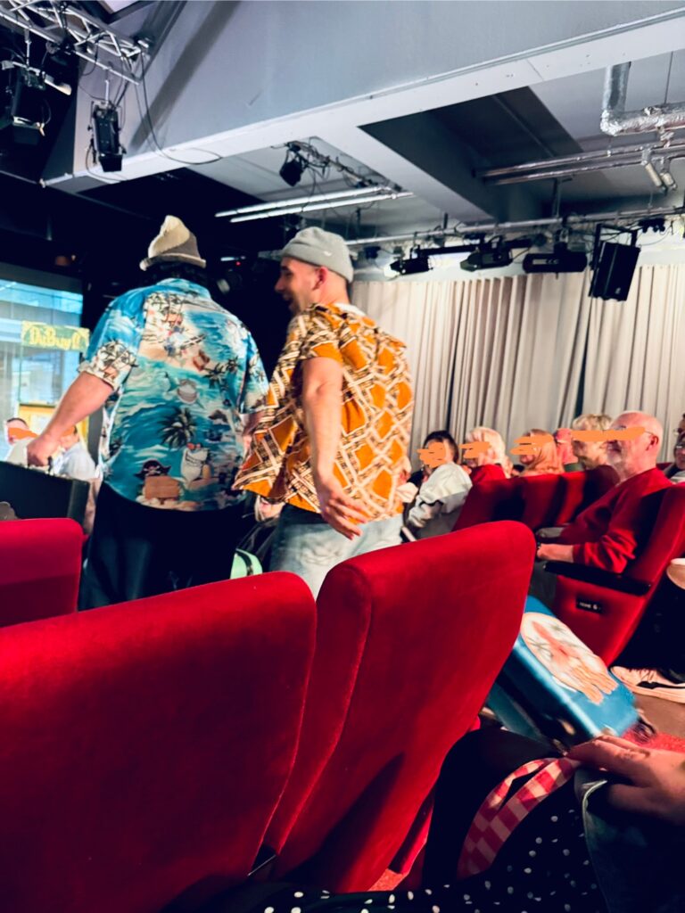 Zwei Schauspieler mit bunten Hemden und Mützen gehen durch den Zuschauerraum eines Theaters mit rotem Stühlen, Publikum und einer Leinwand
