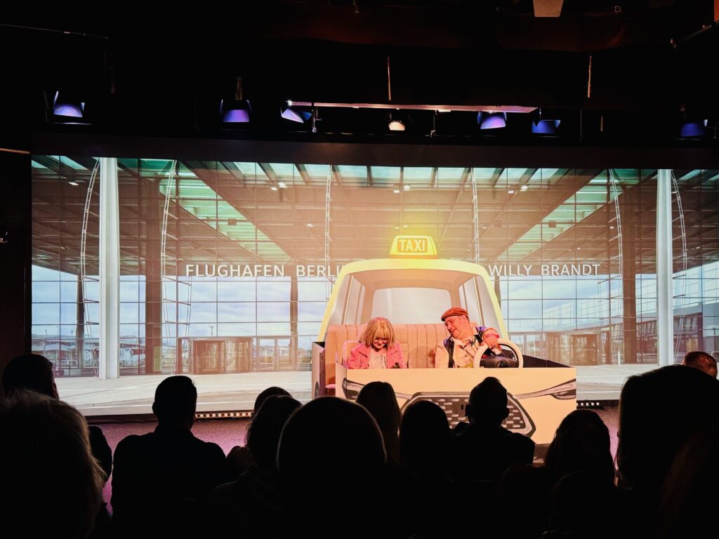 Bühnenbild mit zwei Schauspielern in einem Taxi vor einer Kulisse des Flughafens BER, Publikum im Vordergrund als Silhouetten