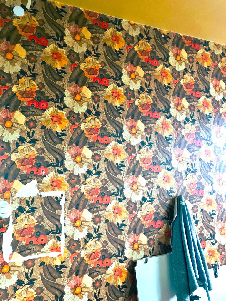 Wand mit altmodischer, angerissener Tapete aus großflächigen Blumenmustern und Blättern, darunter hängt eine Arbeitsjacke an der Wand