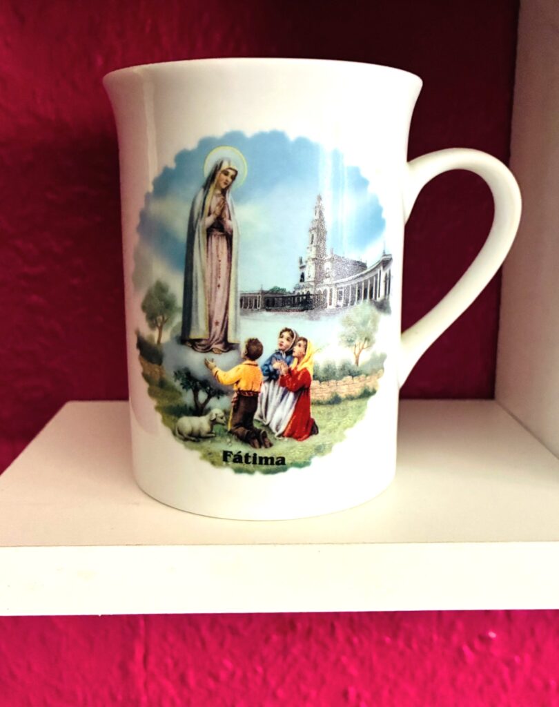 Weiße Tasse mit Illustration der Jungfrau von Fatima mit betenden Kindern auf weißem Regal vor pinkfarbener Wand