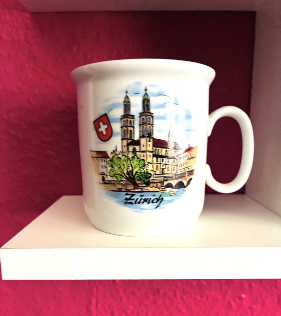 Weiße Tasse mit farbiger Illustration der Zürcher Stadtsilhouette und Schweizer Wappen auf weißem Regal vor pinkfarbener Wand