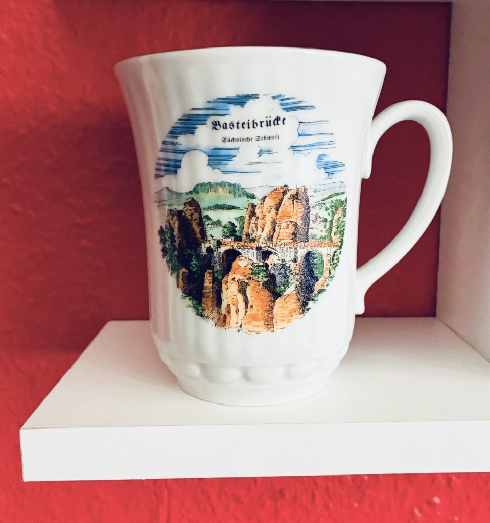 Weiße Tasse mit Illustration der Basteibrücke im Elbsandsteingebirge auf weißem Regal vor pinkfarbener Wand