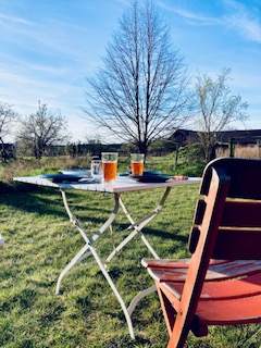 Klappbarer Gartentisch mit zwei Gläsern orangefarbenem Getränk und einem Buch auf einer Wiese neben einem orangen Gartenstuhl unter kahlen Bäumen