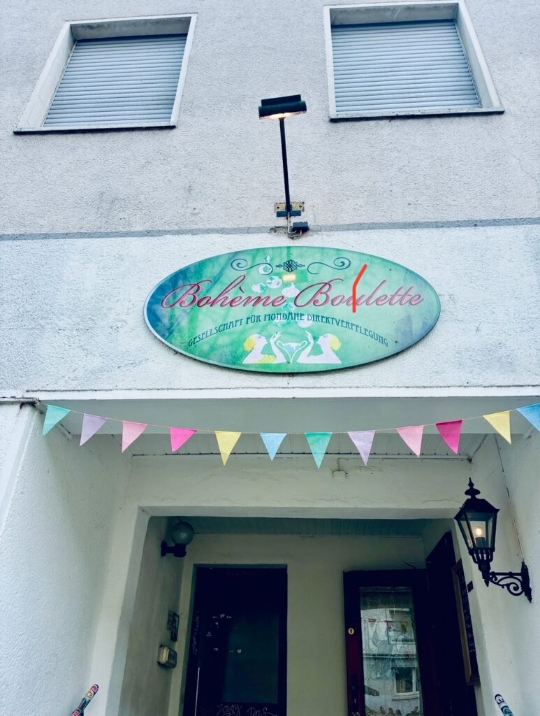 Eingang eines Gebäudes mit ovalem Schild 'Bohème Boulette' und bunter Wimpelkette unter zwei geschlossenen Fenstern