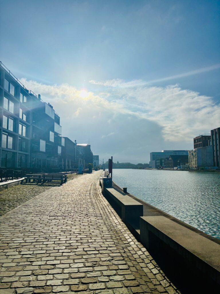 Kopfsteinpflasterweg am Wasser mit modernen Gebäuden links und rechts unter blauem Himmel, Sonne strahlt hinter freundlichen Wolken hervor