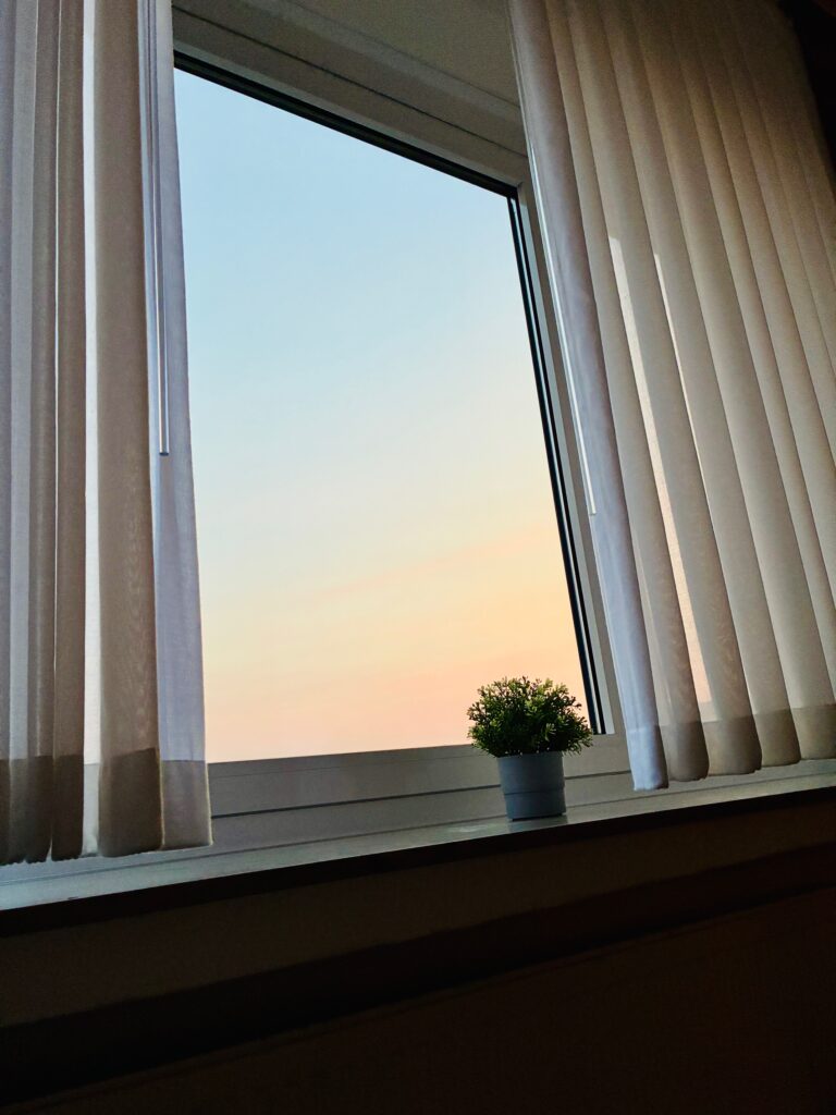 Fenster mit halb geöffneten hellen Vorhängen und kleinem Blumentopf auf der Fensterbank vor pastellfarbenem Himmel