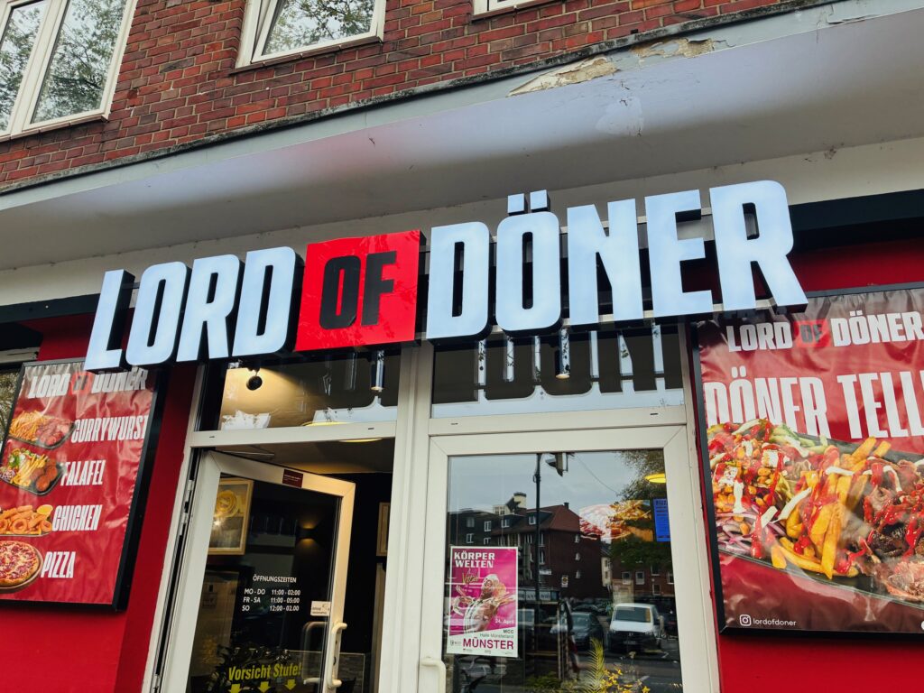 Außenansicht eines Imbiss mit beleuchtetem Schriftzug 'LORD OF DÖNER' über dem Eingang und zwei Werbeplakaten mit Speisen an den Seiten.