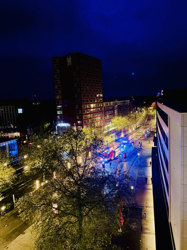 Blick auf eine belebte Straße in der Stadt bei Nacht mit beleuchteten Gebäuden und Bäumen, erleuchtet vom Blaulicht mehrerer Polizeifahrzeuge