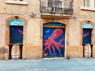 Fassadenwand mit drei blauen Rollläden, auf dem mittleren ein roter Oktopus und auf den seitlichen je einer seiner Arme gemalt, davor zwei Holzfässer