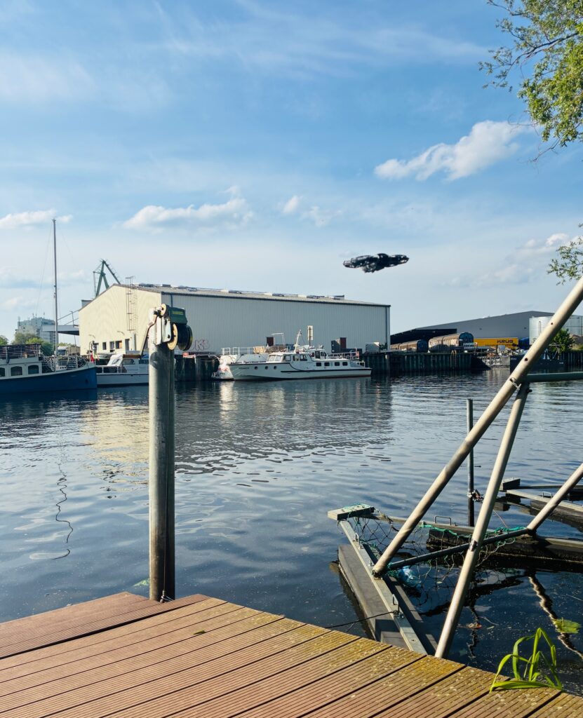 Blick von einem Steg auf einen Fluss mit mehreren Booten und einem großen Lagerhaus im Hintergrund unter blauem Himmel mit Wolken. Vom Steg erhebt sich eine Fotodrohne.