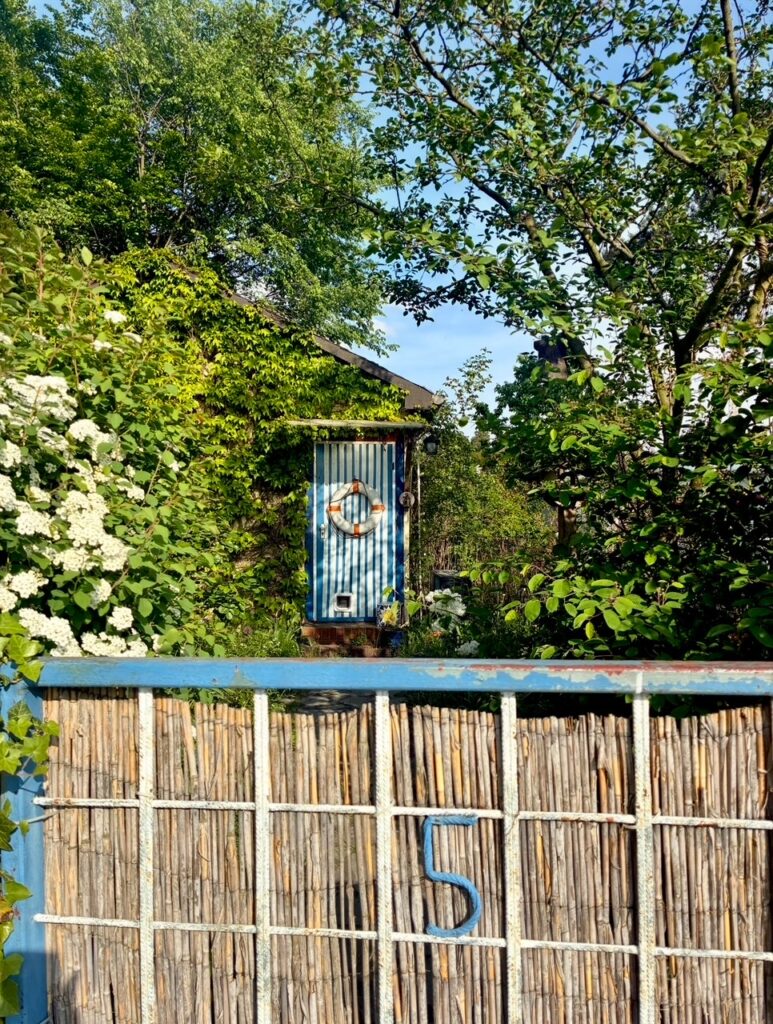 Holztor mit der Zahl 5 vor einem kleinen blau-weiß gestreiften Gartenhäuschen mit Rettungsring an der Tür umgeben von grünem Laub und blühenden Sträuchern