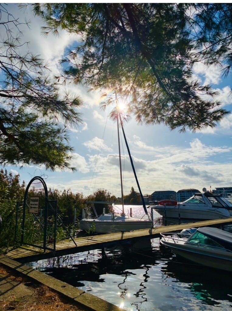 Segelboot und Motorboote liegen an einem Steg in einem Hafen unter Bäumen bei Sonnenschein unter Himmel mit freundlichen Wolken