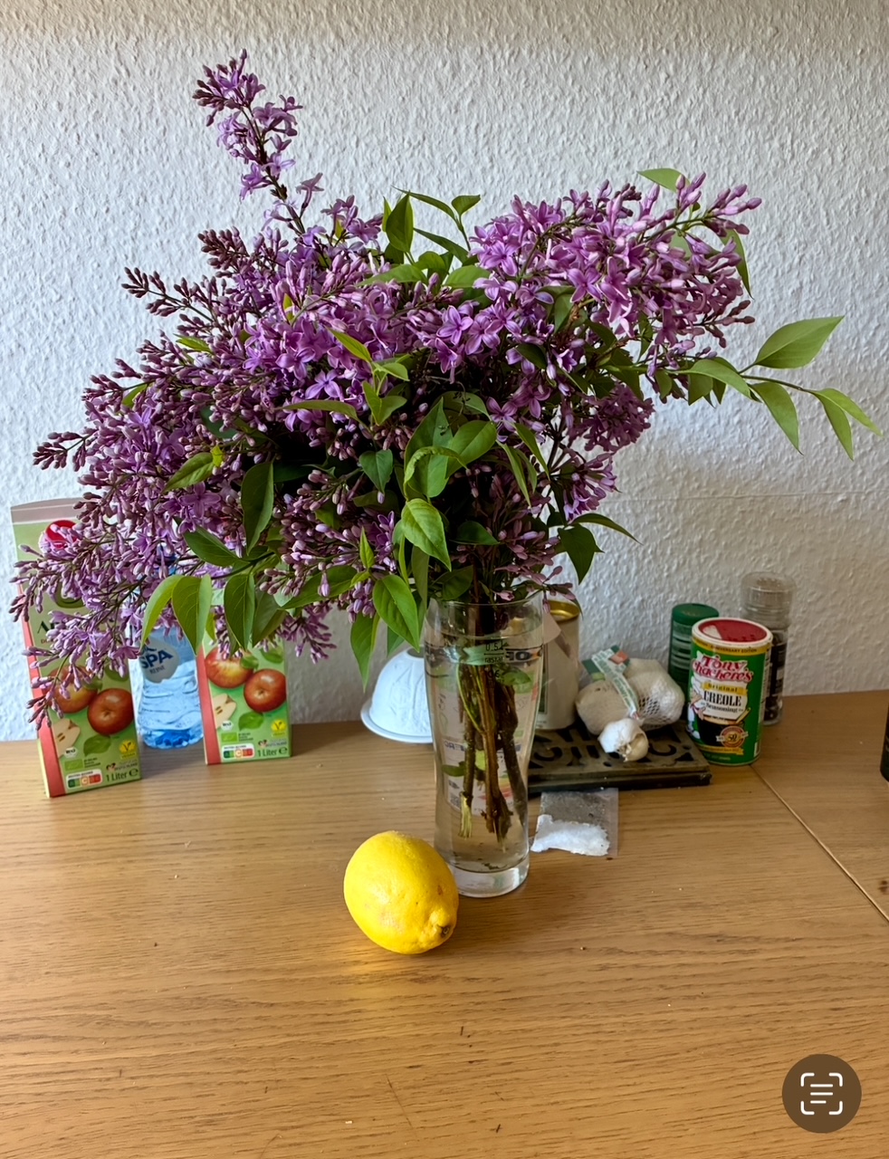 Großer Strauß lila Flieder mit grünen Blättern in einem Glas auf einem Holztisch mit einer Zitrone davor und verschiedenen Lebensmitteln im Hintergrund