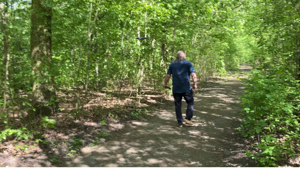 Ein Mann in blauer Kleidung geht auf einem von Bäumen gesäumten Waldweg. Hinter ihm fliegt eine Drohne