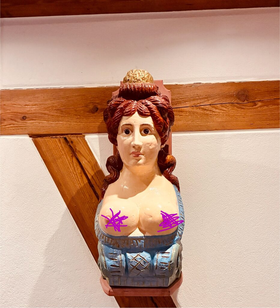 Galionsfigur aus Holz in Form einer Frau mit hochgestecktem Haar und blauem Kleid an einer weißen Wand mit sichtbarem Holzbalken