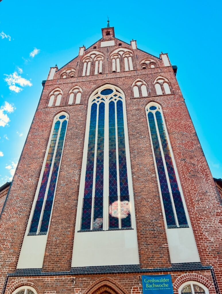 Fassade eines hohen Backsteingebäudes mit großen, schmalen Buntglasfenstern und blauem Himmel im Hintergrund