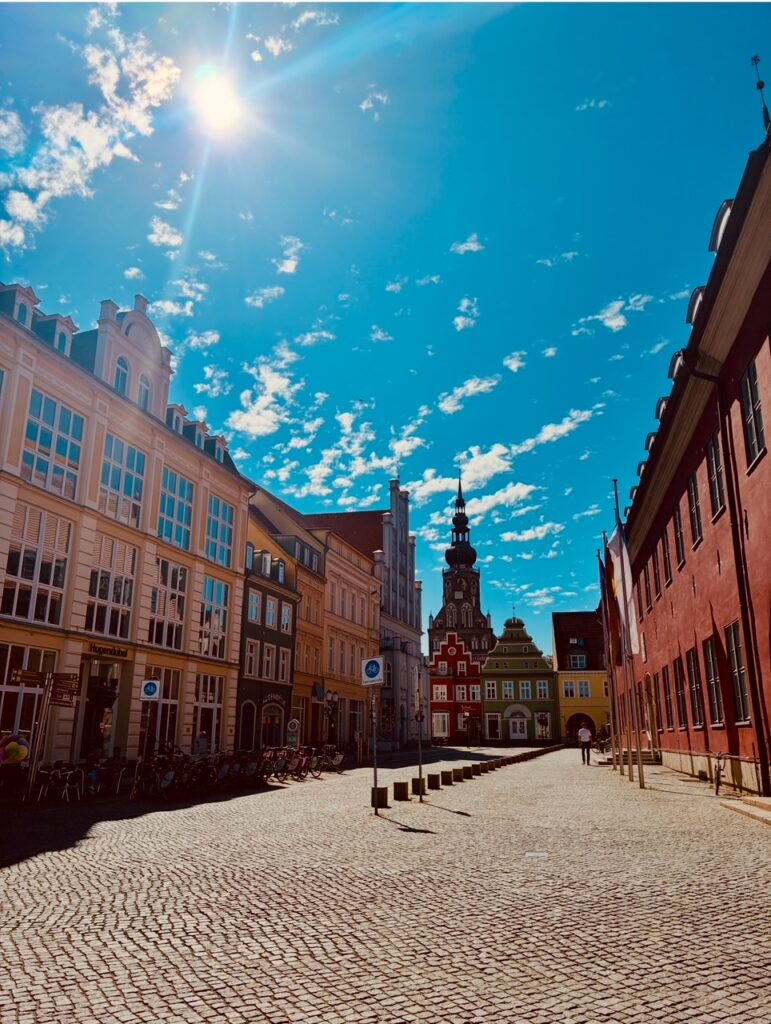 Sonniger Himmel über gepflastertem Platz mit historischen Gebäuden und Kirchturm im Hintergrund