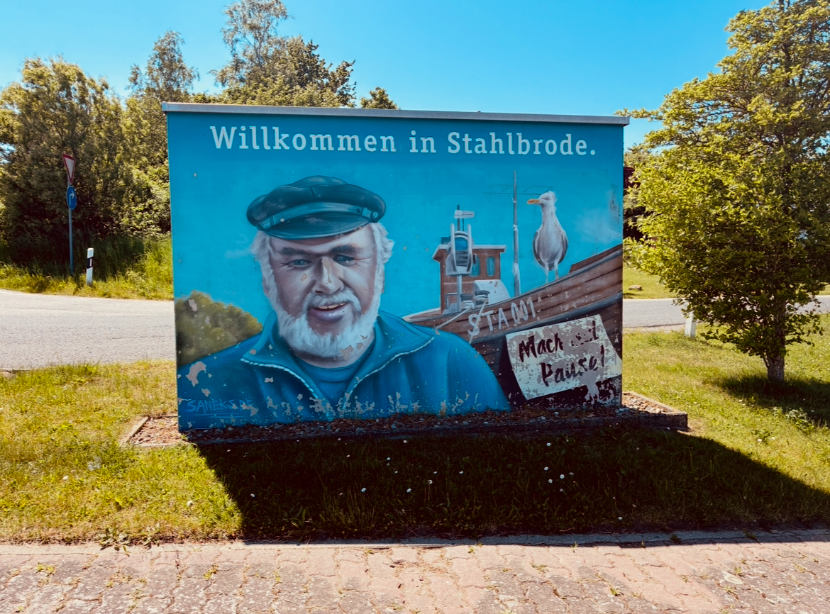 Wandbild mit der Aufschrift 'Willkommen in Stahlbrode' zeigt einen Mann mit Mütze, ein Schiff und eine Möwe sowie ein Schild mit der Aufschrift 'Mach Pause!'