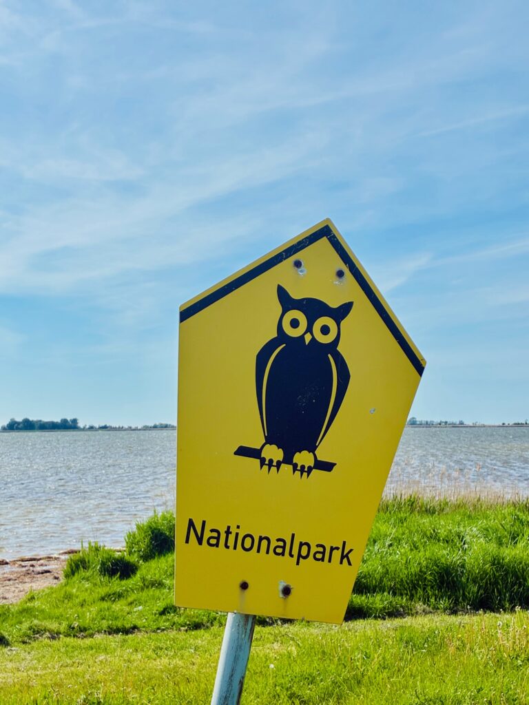 Gelbes Verkehrsschild mit schwarzer Eule und der Aufschrift 'Nationalpark' vor einer Wasserfläche und grünem Ufer