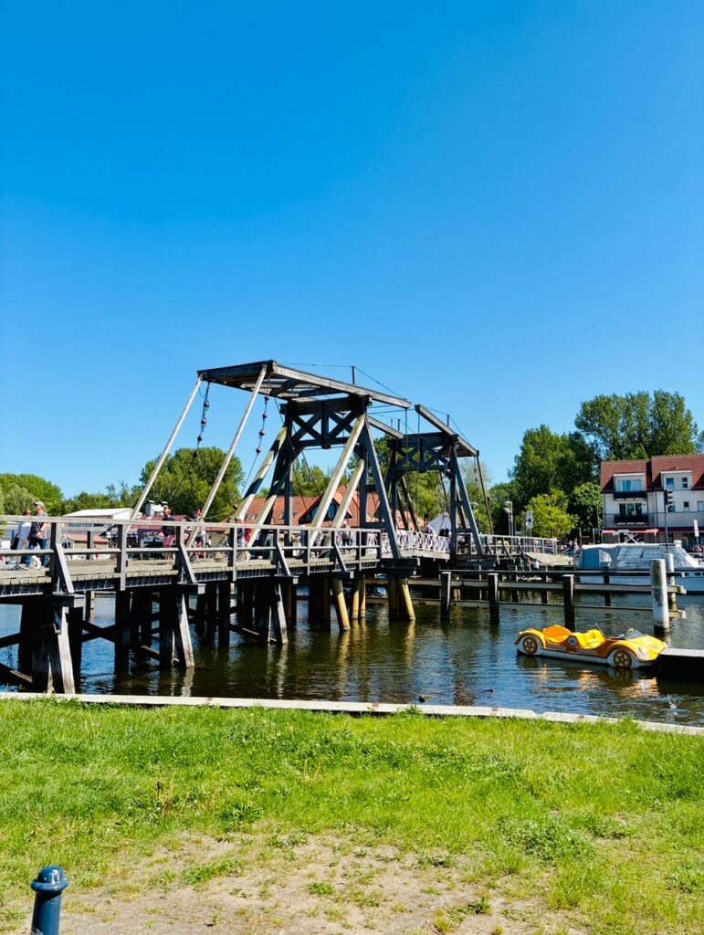 Holzbrücke mit Fachwerk über einem Fluss, daneben ein gelbes Tretboot im Wasser und im Hintergrund Häuser und Bäume bei klarem Himmel