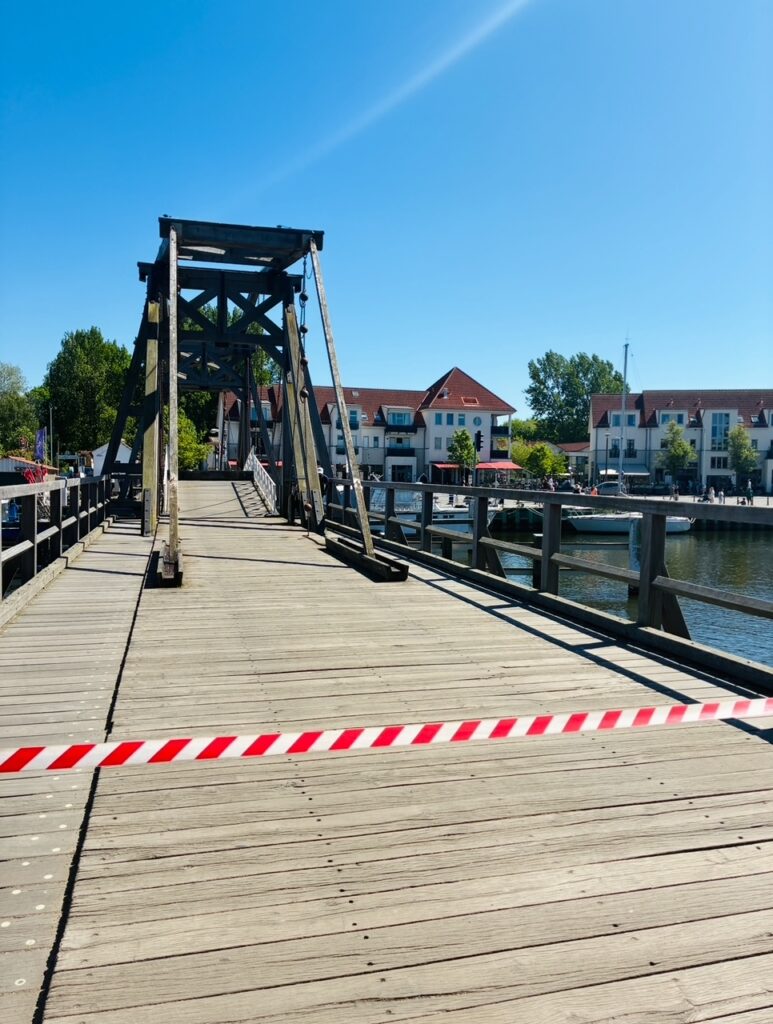 Holzbrücke mit Absperrband vor dem Zugang und Wohnhäusern am Ufer eines Flusses im Hintergrund