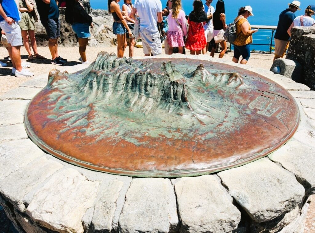 Bronzefläche mit erhabener Reliefdarstellung des Tafelgebirges auf einem steinernen Podest vor einer Gruppe von Menschen mit blauem Himmel