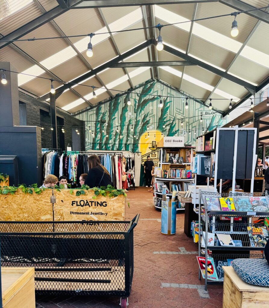 nnenansicht eines Markthallenraums mit Verkaufsständen für Kleidung, Schmuck und Bücher unter einem Glasdach mit sichtbaren Stahlträgern